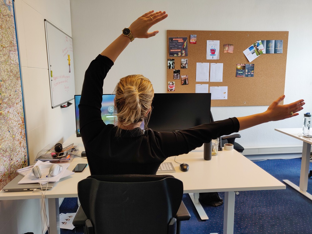 Foto van medewerker die de oefening genaamd wave uitvoert achter haar bureau. Ze steekt haar armen omhoog en beweegt met beide armen naar één kant.